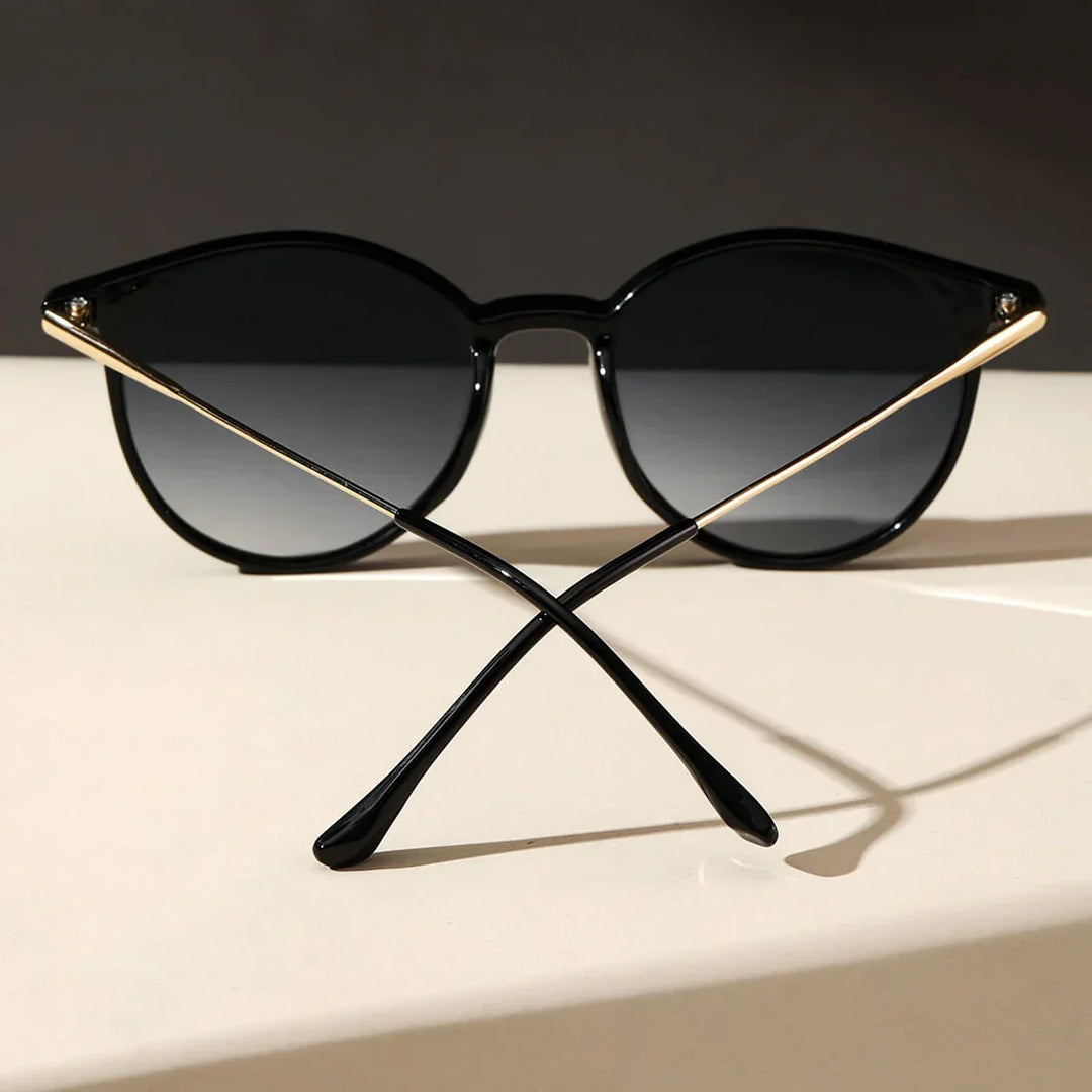 A pair of black sunglasses with round frames, featuring UV protection, is placed on a light surface and photographed from the front with the arms crossed in an X shape. The dark background creates a striking contrast.
