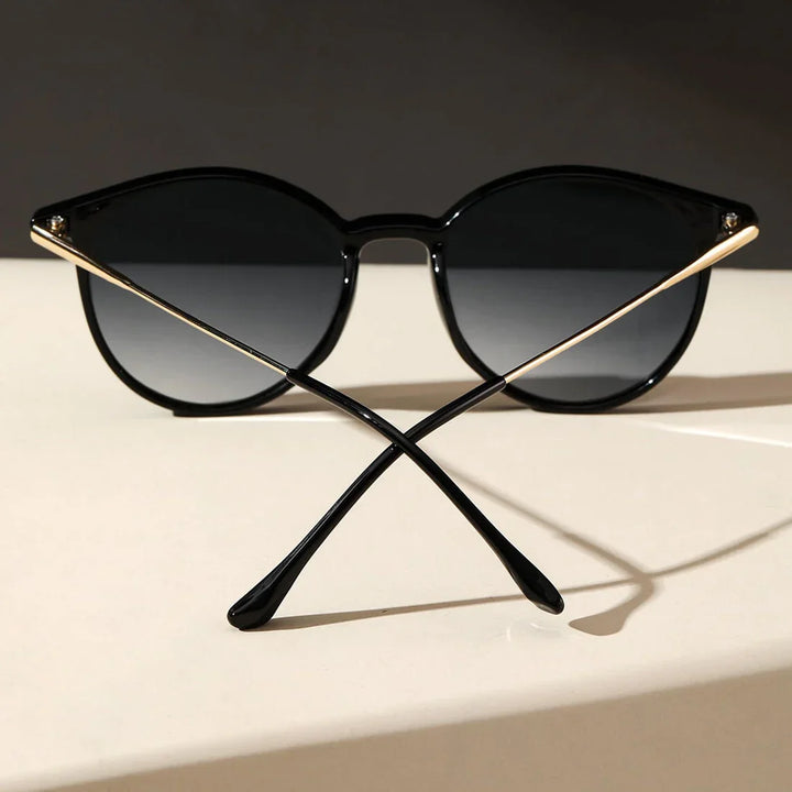 A pair of black sunglasses with round frames, featuring UV protection, is placed on a light surface and photographed from the front with the arms crossed in an X shape. The dark background creates a striking contrast.