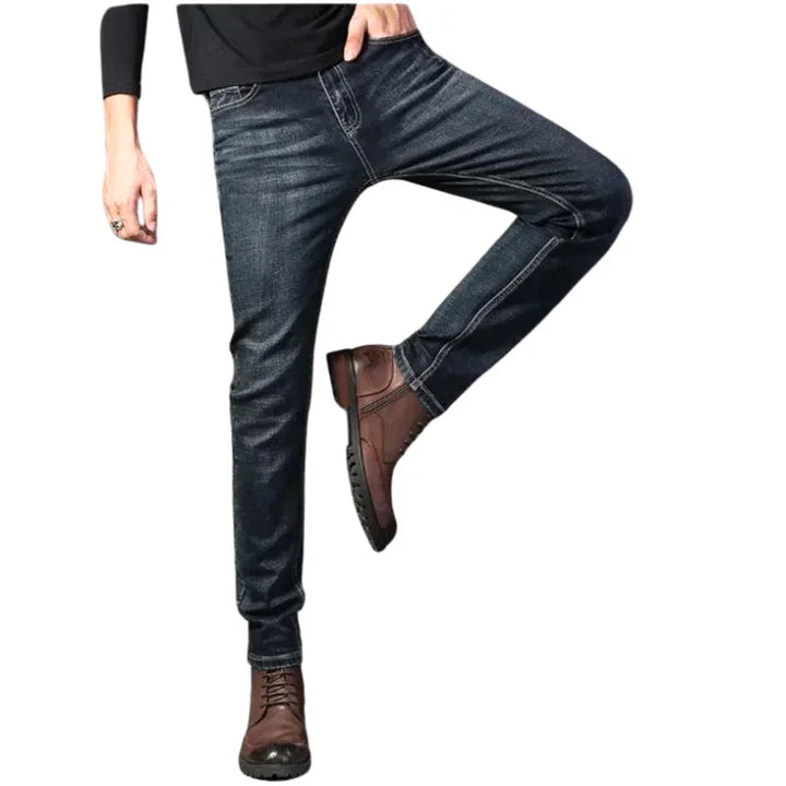 A person wearing dark blue slim-fit jeans, a black long-sleeve shirt, and brown lace-up boots poses with one leg bent and the other straight, against a white background.