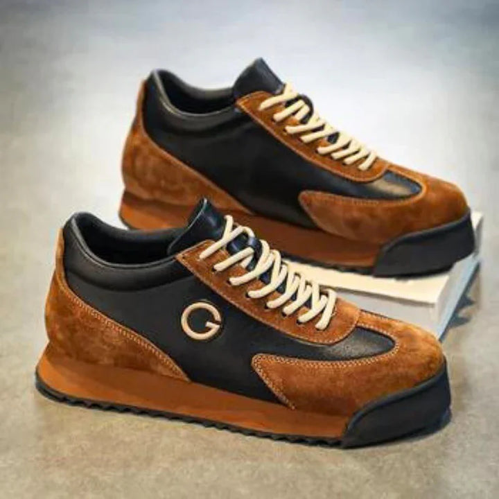 A pair of sleek sneakers with luxe leather suede, brown and black accents, white laces, and a circular G logo on the side, displayed on a grey surface with one shoe elevated on a box.