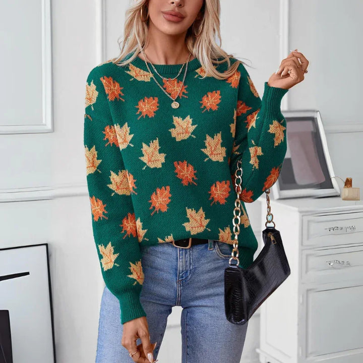 A woman wearing a comfortable sweater in soft knit material, featuring orange and yellow maple leaf patterns, light blue jeans, and holding a black purse with a chain strap, stands in a modern, white room.