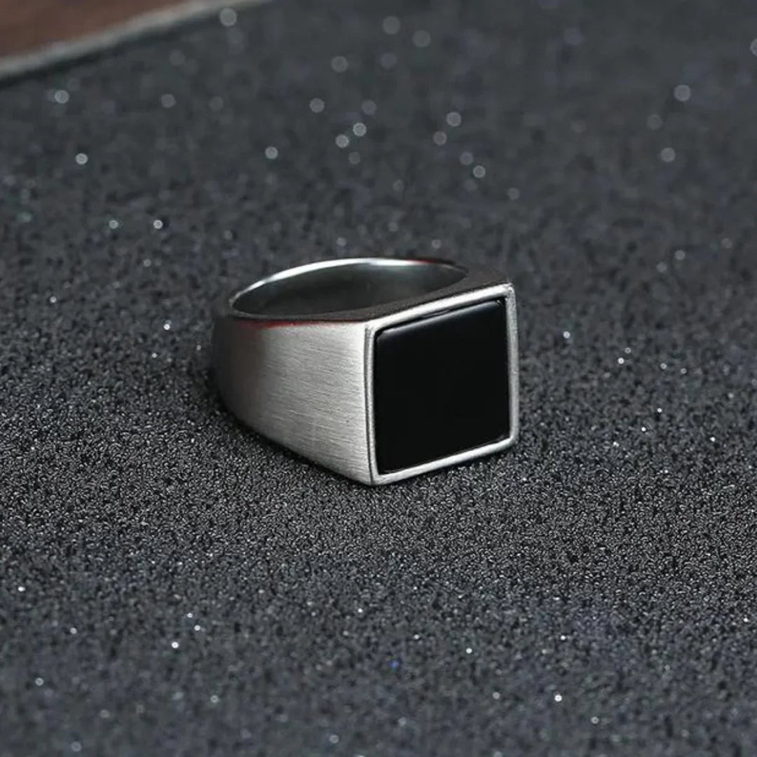A silver ring with a large, flat, black rectangular face and a polished finish rests on a textured, dark surface sprinkled with fine glitter.