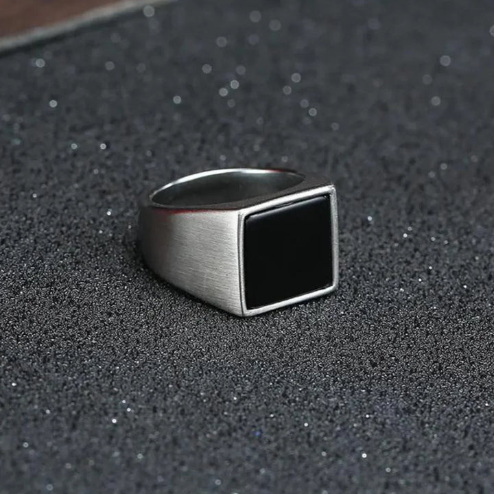 A silver ring with a large, flat, black rectangular face and a polished finish rests on a textured, dark surface sprinkled with fine glitter.