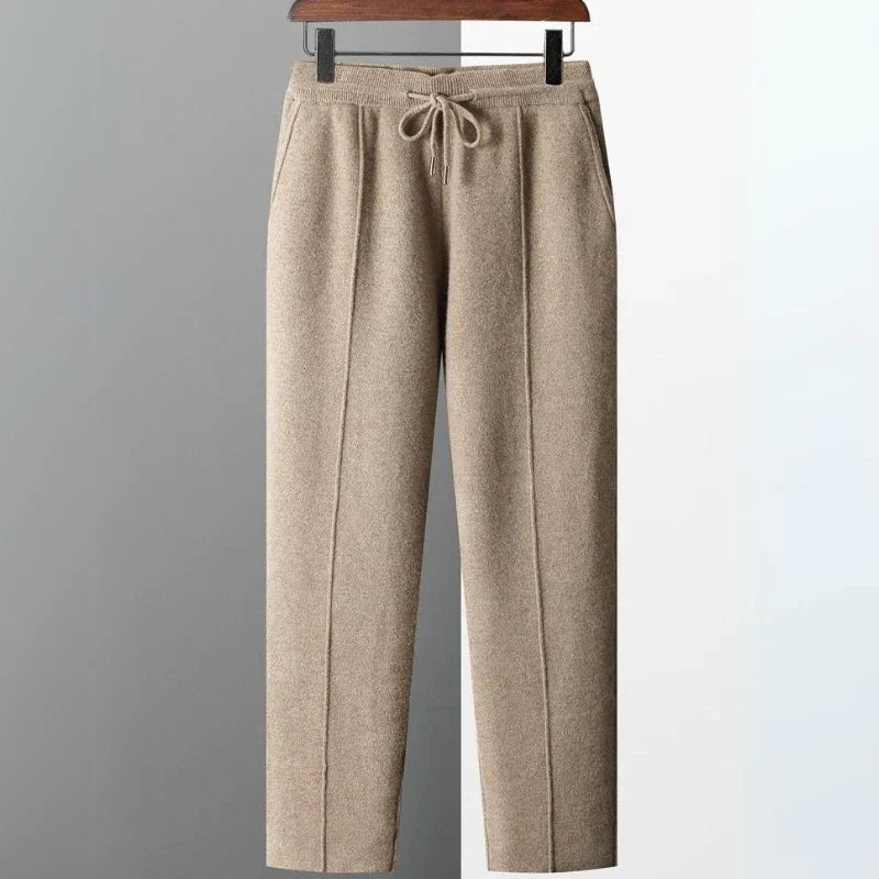 Beige Merino wool pants with a relaxed fit hang on a wooden hanger against a light grey and white background. These comfortable pants feature side pockets and durable construction for everyday wear.