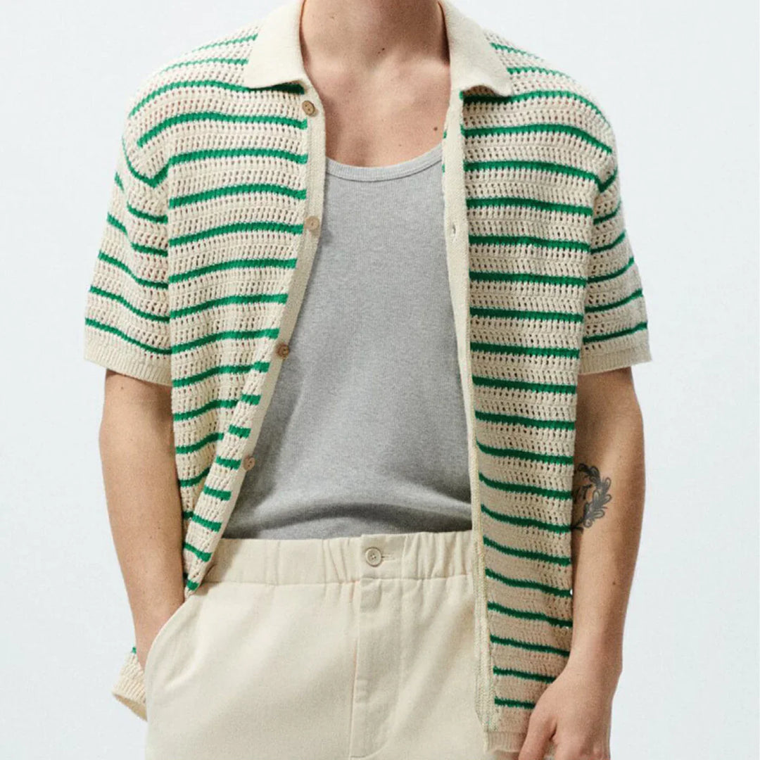 A person wearing a beige and green striped short-sleeve retro knit polo cardigan over a light gray T-shirt, paired with high-waisted beige pants; hands are in pockets and a tattoo is visible on one arm.
