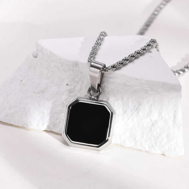 A silver necklace with a square Onyx Stone Amulet pendant rests on a white, textured stone surface. The chain is rope-style, and the collectable jewelry piece features a shiny metal frame with beveled edges.