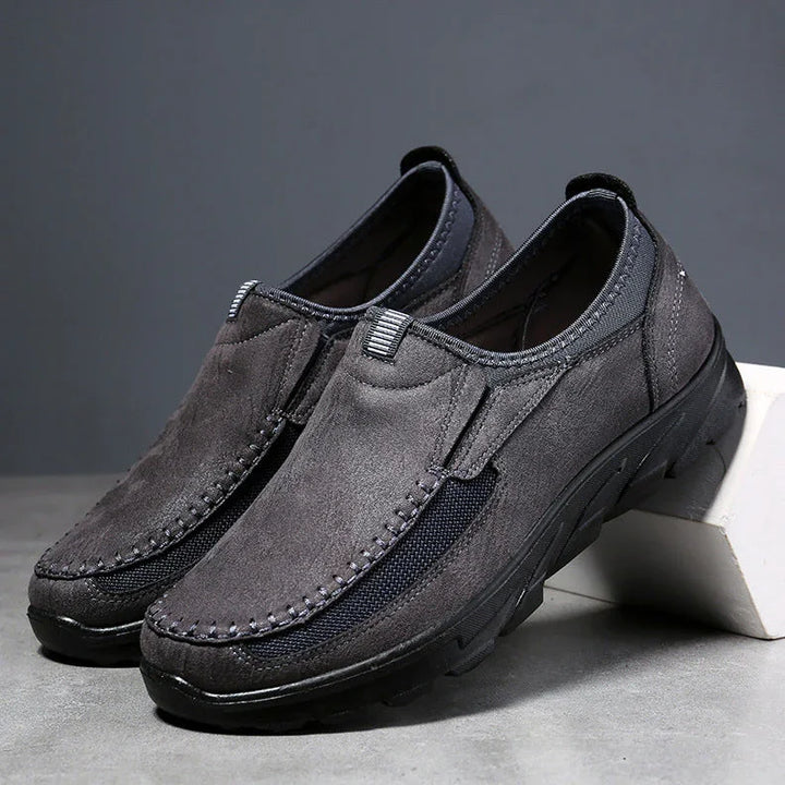 A pair of gray suede breathable loafers with black mesh side panels and thick black rubber soles, offering a comfortable fit. Displayed on a gray surface, one shoe is slightly elevated on a white block.
