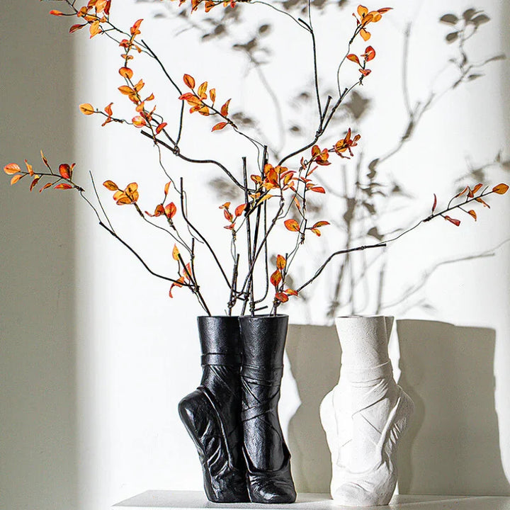 Two ballet-inspired vases shaped like boots—one black, one white—sit on a surface with orange-leaved branches in the black ceramic vase, casting artistic shadows on the wall. Perfect for a touch of contemporary chic décor.