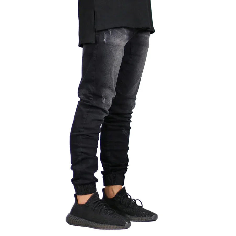 A person wearing black straight tapered leg jeans and black sneakers stands against a white background. The image shows only the legs and lower torso, with the upper body not visible.