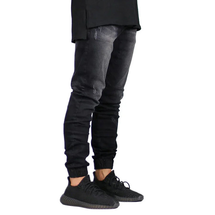 A person wearing black straight tapered leg jeans and black sneakers stands against a white background. The image shows only the legs and lower torso, with the upper body not visible.