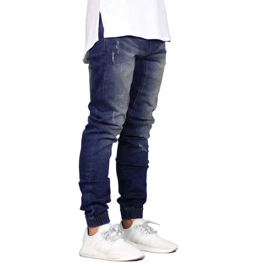 A person wearing weathered denim jeans with a straight tapered leg, a long white shirt, and white sneakers stands against a white background. Only the lower half of the body is visible, creating a subtle vintage aesthetic.