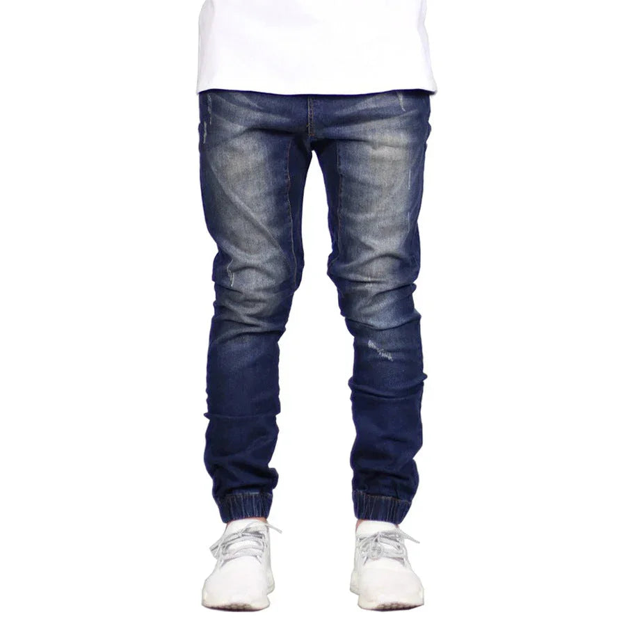 A person wearing dark blue, straight tapered leg, jogger-style jeans with faded details, paired with a white shirt and white sneakers. Only the lower half of the body is visible, giving off a subtle vintage aesthetic.