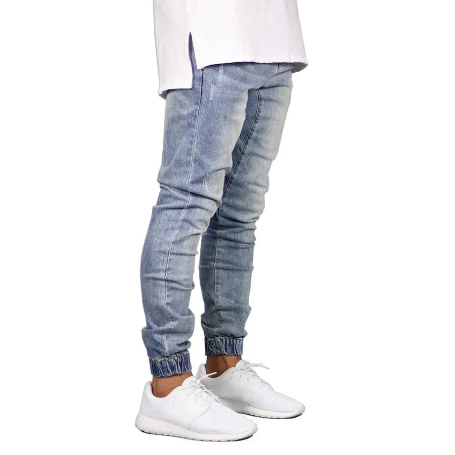 A person wearing light blue jogger jeans with elastic cuffs, a white shirt with side slits, and white athletic sneakers. The straight tapered leg design adds a modern twist to the vintage aesthetic against a clean white background.