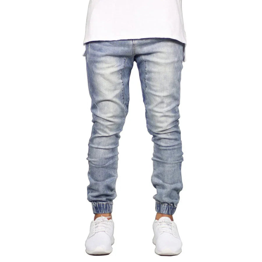 A person wearing light blue straight tapered leg jogger jeans, a white t-shirt, and white sneakers stands against a plain white background. The photo is cropped at the shoulders.