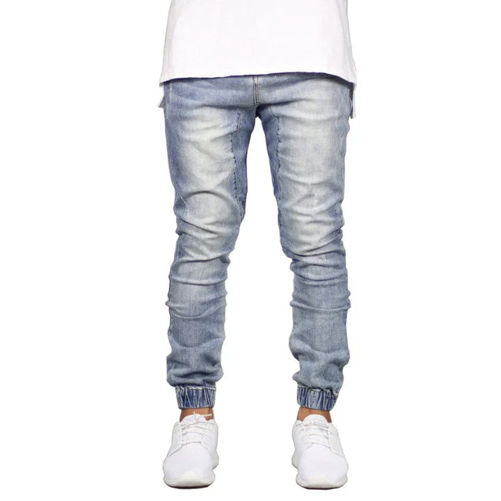 A person wearing light blue straight tapered leg jogger jeans, a white t-shirt, and white sneakers stands against a plain white background. The photo is cropped at the shoulders.