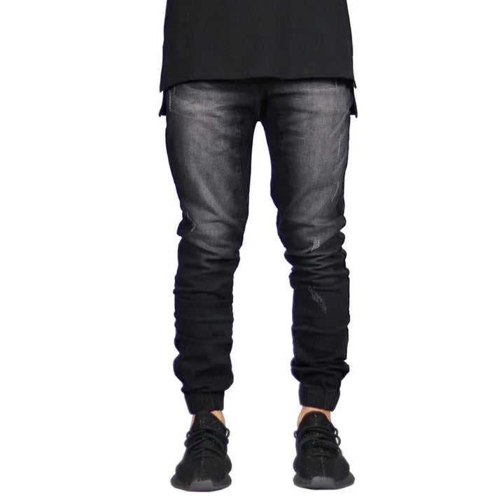 A person wears black jogger jeans with a faded effect and straight tapered leg, paired with a long black shirt and black sneakers. The image is cropped above the shoulders, highlighting a modern look with vintage aesthetic.