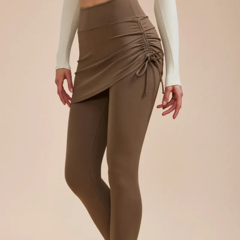 A person wearing high-waisted leggings with a matching ruched skirt overlay and a fitted cream top, perfect for an autumn wardrobe, stands against a plain beige background.