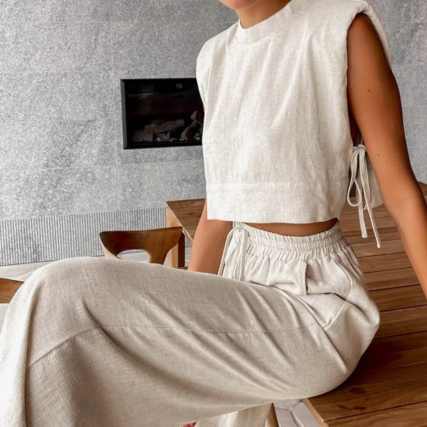 A person wearing a sleeveless, light beige Belgian Linen Blend cropped top and matching wide-leg pants is seated indoors near a wooden table and chair, with a fireplace and stone wall in the background.