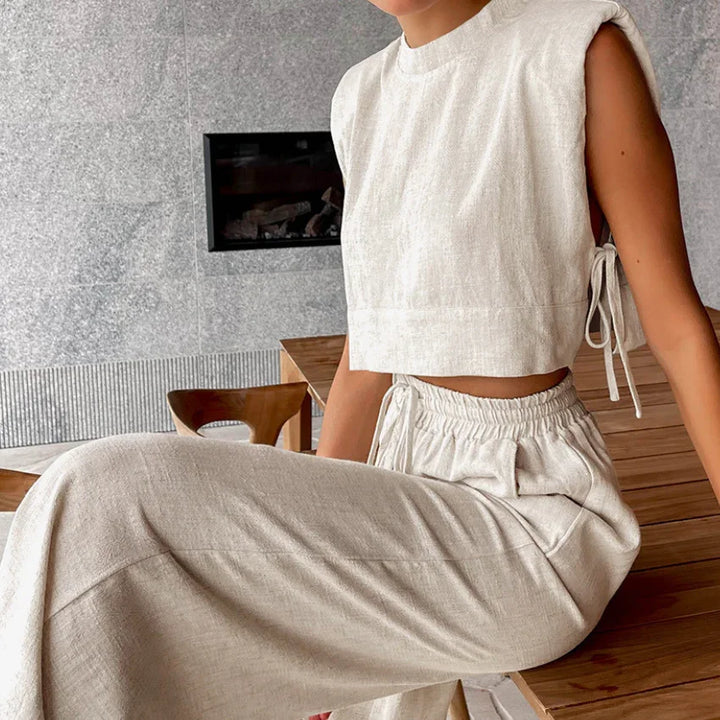 A person wearing a sleeveless, light beige Belgian Linen Blend cropped top and matching wide-leg pants is seated indoors near a wooden table and chair, with a fireplace and stone wall in the background.