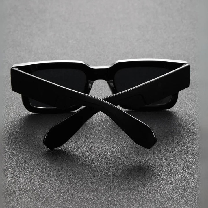 A pair of black lightweight sunglasses with thick frames is placed upside down on a dark textured surface, with the arms crossed in an X shape in front of the lenses.
