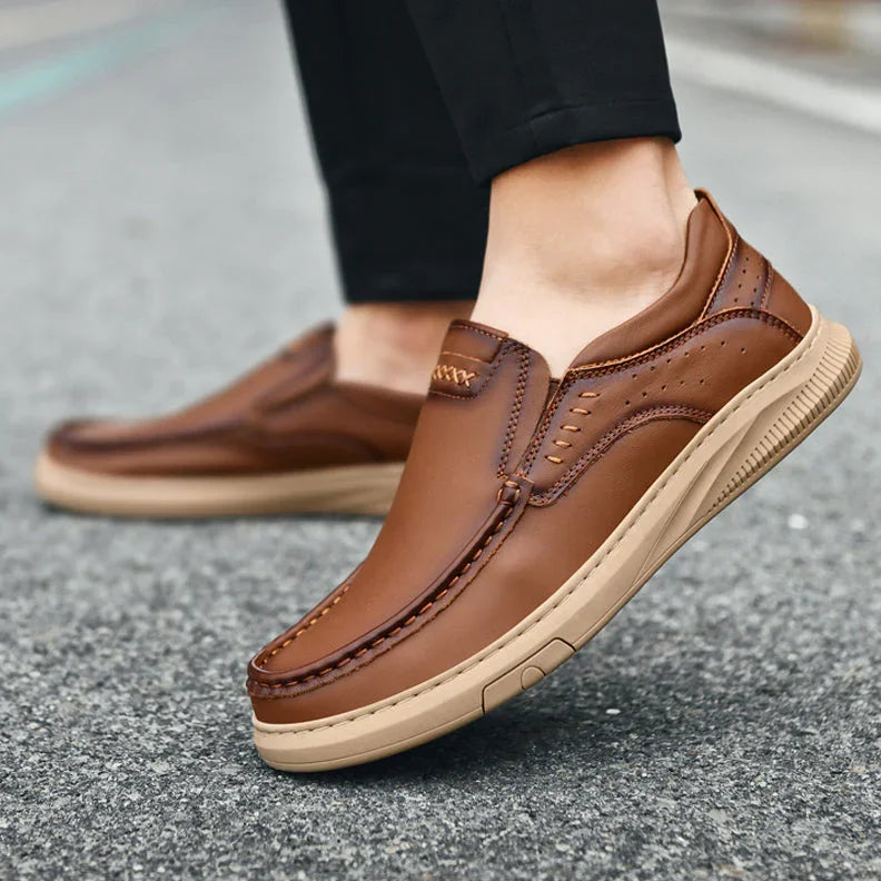 A person wearing men's cowhide leather slip-on shoes with tan soft soles stands on gray pavement. The shoes feature visible stitching and perforated detailing, paired with black pants.