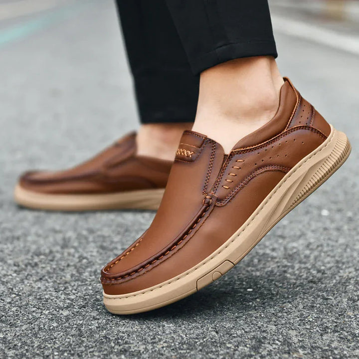 A person wearing men's cowhide leather slip-on shoes with tan soft soles stands on gray pavement. The shoes feature visible stitching and perforated detailing, paired with black pants.