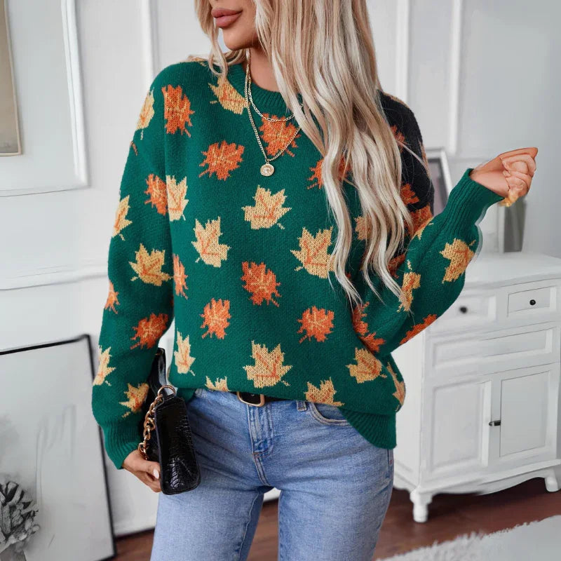 A woman with long blonde hair wears a comfortable sweater in soft knit material, featuring a fall maple leaf pattern in orange and yellow, blue jeans, and holds a black purse. She stands indoors in a modern, white room.