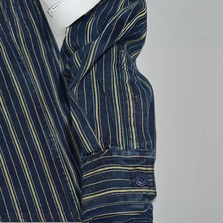 A close-up of a relaxed-fit button-down navy shirt sleeve with thin yellow and white vertical stripes, showing a buttoned cuff. The sleeve is partially folded, and a white object is tucked under the arm.