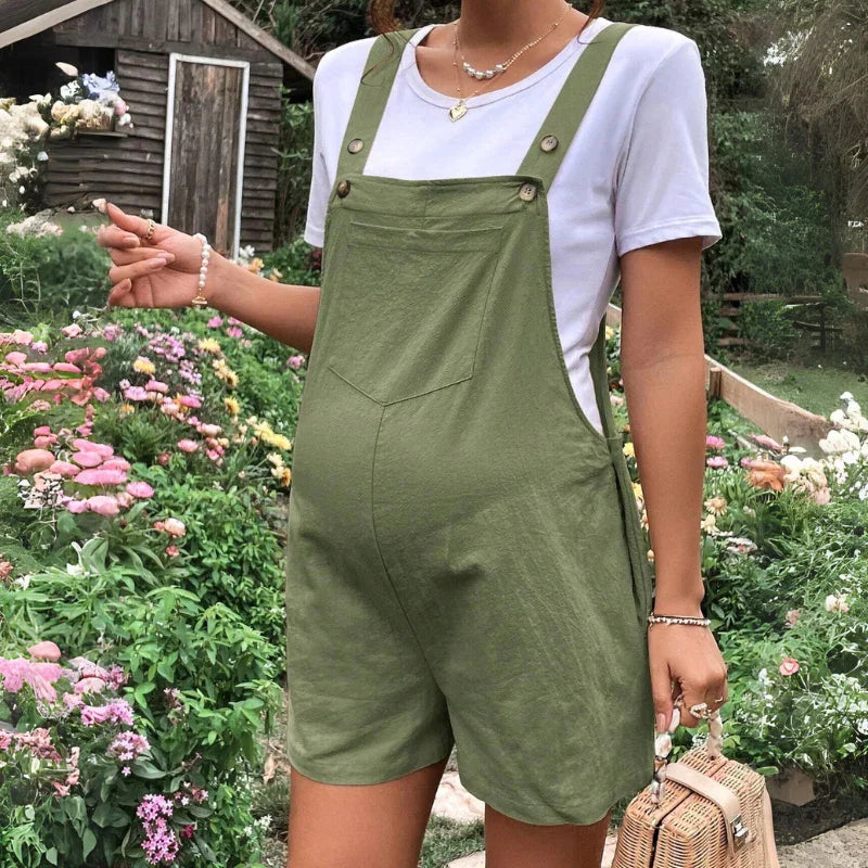 A person wearing comfortable maternity overalls and a white t-shirt stands in a garden filled with colorful flowers, holding a woven handbag. A wooden shed is visible in the background.