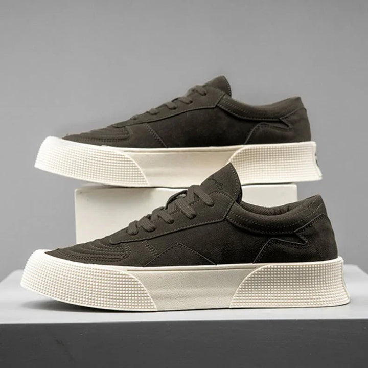 A pair of dark brown suede lace-up sneakers with thick, textured white soles and comfortable cushioning are displayed on a gray and white platform against a plain gray background.