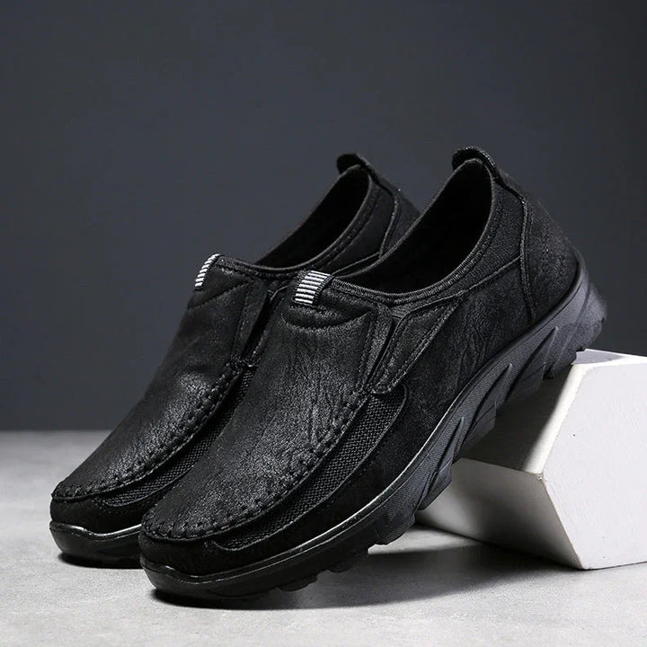 A pair of black Men Leather Casual Shoes with textured soles, displayed on a gray surface. One shoe is propped against a white block, set against a dark gray background—designed for a comfortable fit and effortless style.
