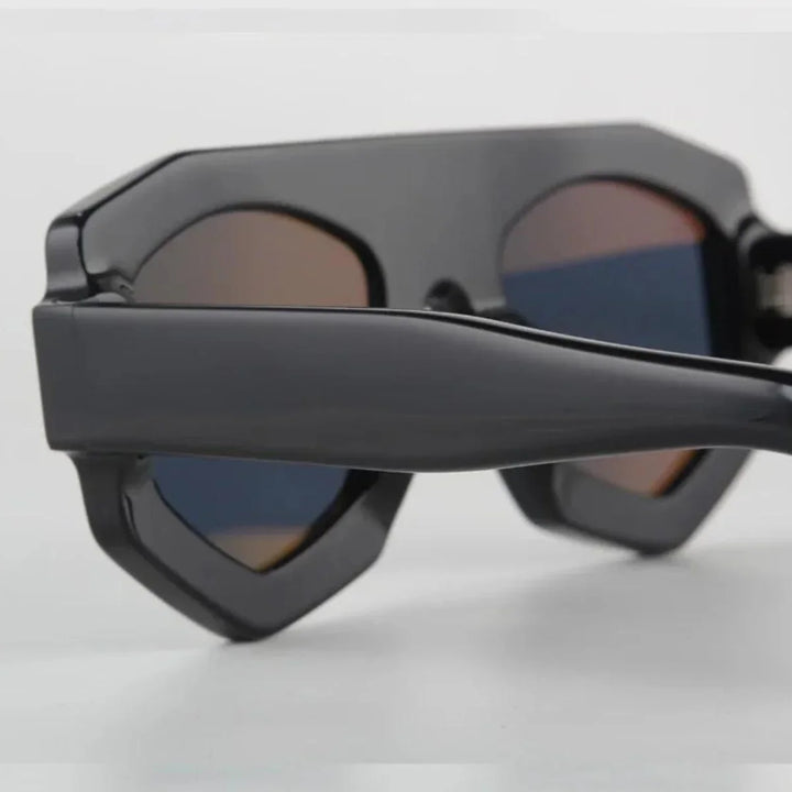 Close-up of black acetate geometric sunglasses with thick frames and dark, slightly reflective lenses, offering UV400 protection while resting on a white surface.