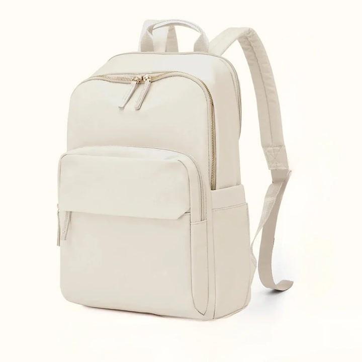 A minimalist, light beige, water-resistant backpack with gold zippers, a large front pocket, side pockets, adjustable padded shoulder straps, and a dedicated laptop compartment, displayed against a plain white background.