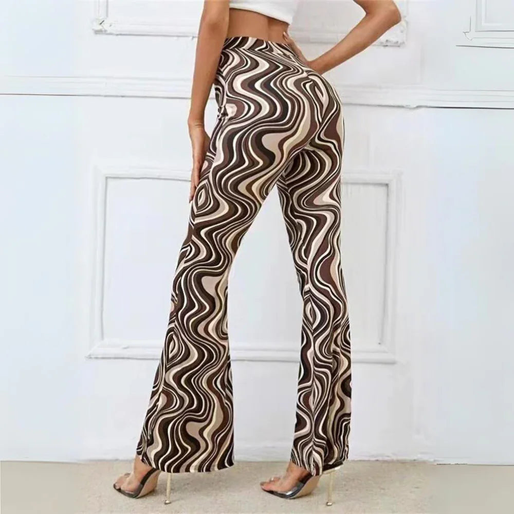 A person stands with their back to the camera, wearing trendy statement pants featuring a brown, beige, and black wavy swirl pattern and a white crop top, posing in high-heeled clear shoes against a white wall.