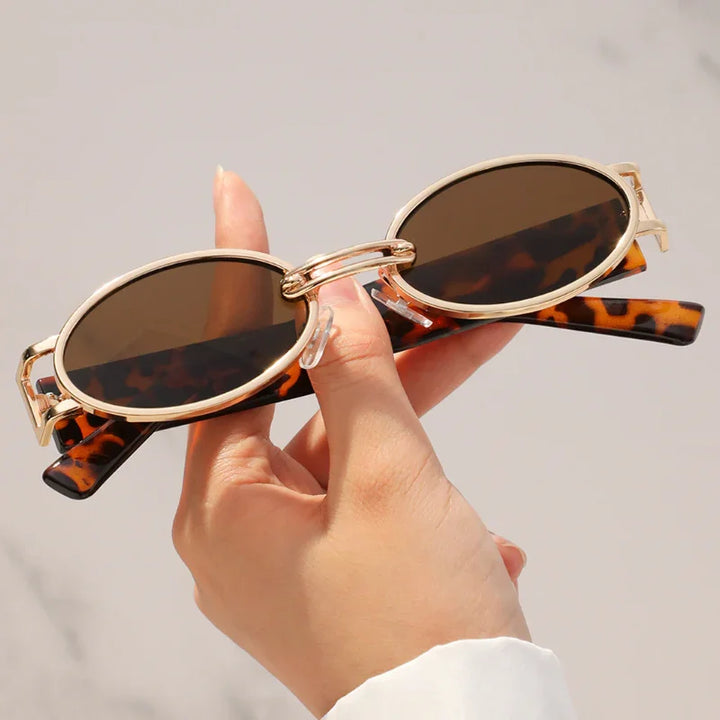 A hand holds retro oval sunglasses with brown lenses and a durable metal frame. The gold temples feature a brown tortoiseshell pattern, while the softly blurred, light background highlights their stylish UV400 protection.