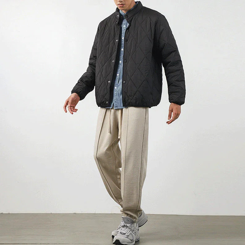 A person stands against a plain background wearing a black diamond-stitched padded jacket over a blue button-up shirt, beige loose-fitting trousers, and gray sneakers. Their face is partially out of frame.