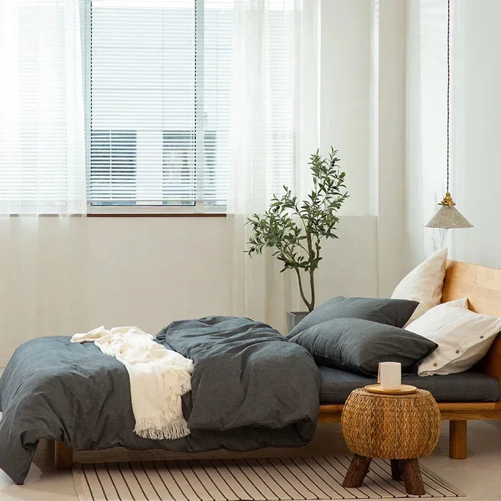 Minimalist bedroom with gray bedding, wooden bed, indoor plant, and woven nightstand from Rovelli