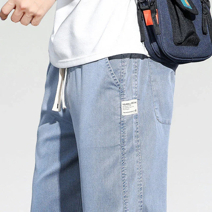 A person wearing Summer Loose Men Jeans in light blue with a white shirt and navy crossbody bag. Only the lower torso and part of one arm are visible against a plain light background.