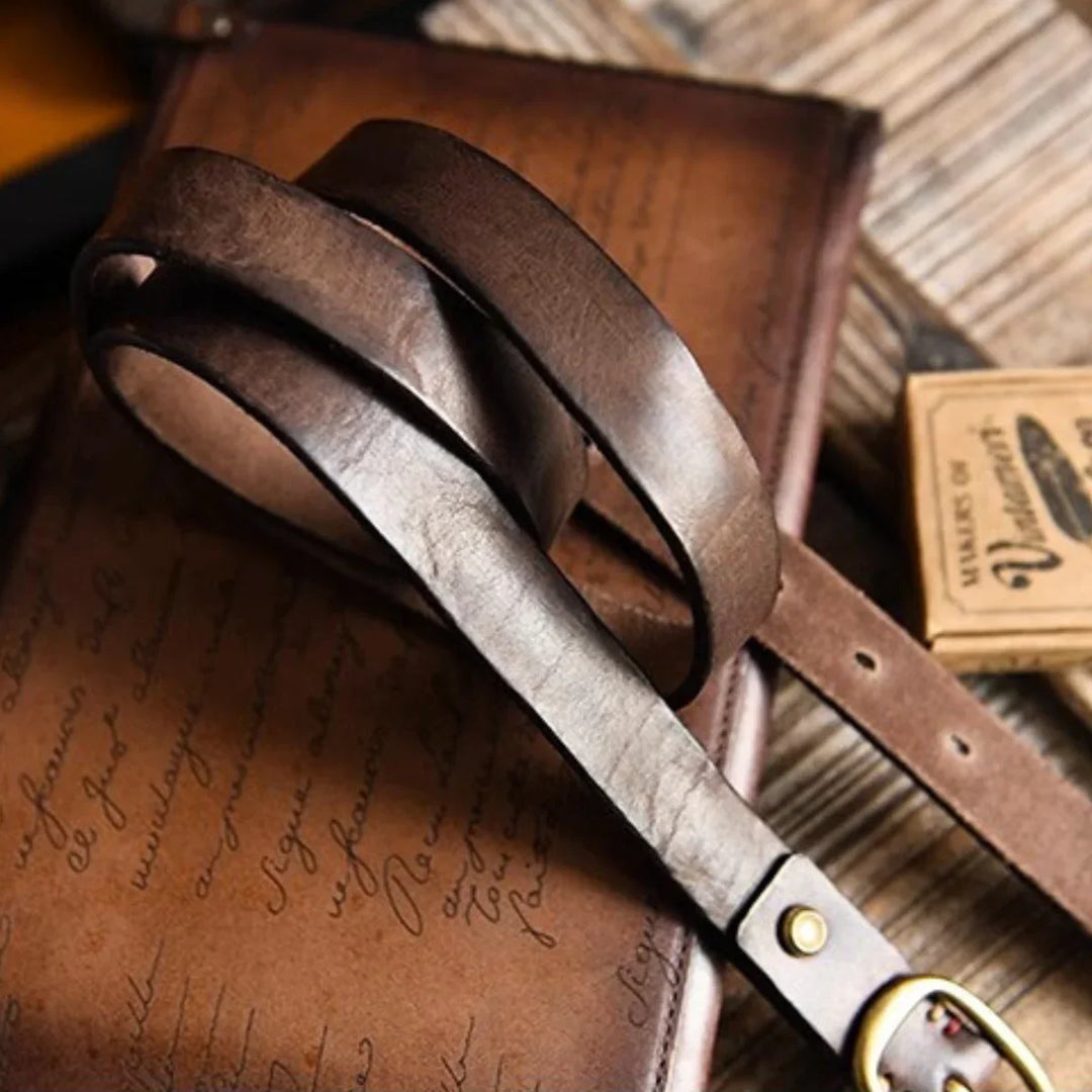 A close-up of a brown leather belt with brass buckle, lying on top of a vintage leather-bound journal with cursive writing, evokes timeless country fashion on a rustic wooden surface.