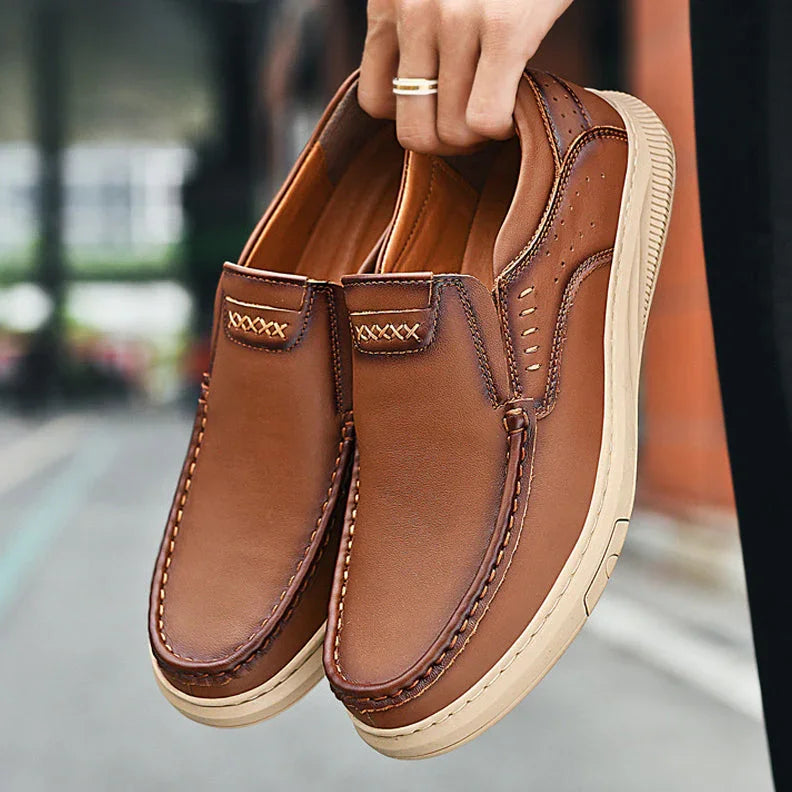 A person holds a pair of stylish Men's Cowhide Leather Shoes with soft soles and decorative stitching. These slip-on brown loafers feature beige rubber soles, displayed outdoors.