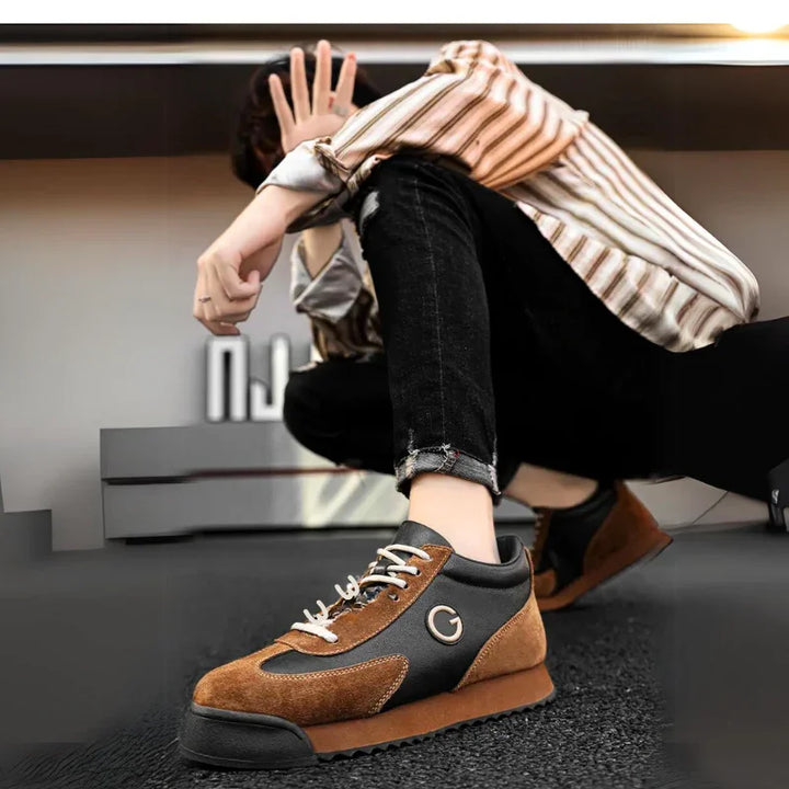 A person in a striped shirt and ripped black jeans sits on the ground, holding a hand up to partially cover their face. The focus is on their luxe leather suede sneakers with white laces and a G logo.