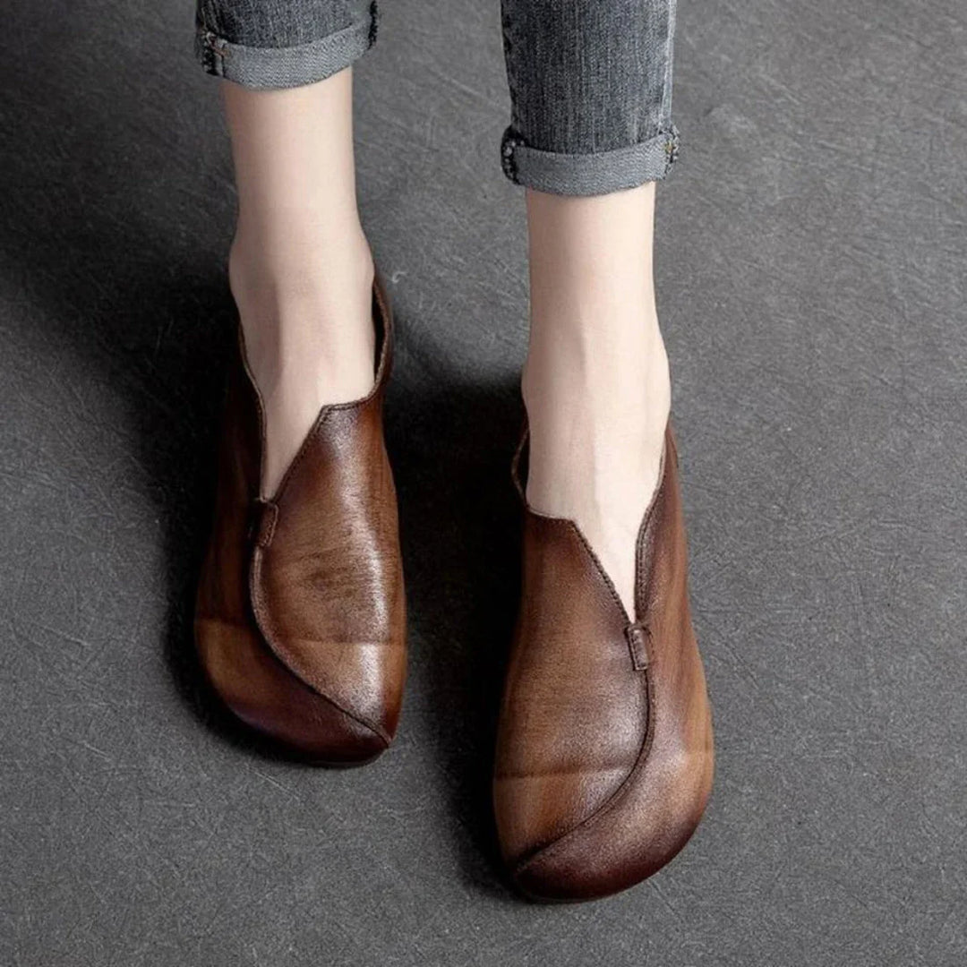 Person wearing vintage-style brown leather shoes with a unique, asymmetrical design and cushioned insole, standing on a textured dark gray surface. Their jeans are rolled up just above the ankles.