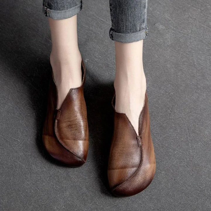 Person wearing vintage-style brown leather shoes with a unique, asymmetrical design and cushioned insole, standing on a textured dark gray surface. Their jeans are rolled up just above the ankles.