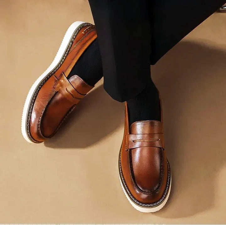 A person wearing black pants and black socks is seated with feet crossed, sporting business casual brown Italian leather penny loafers with white soles on a light brown floor.