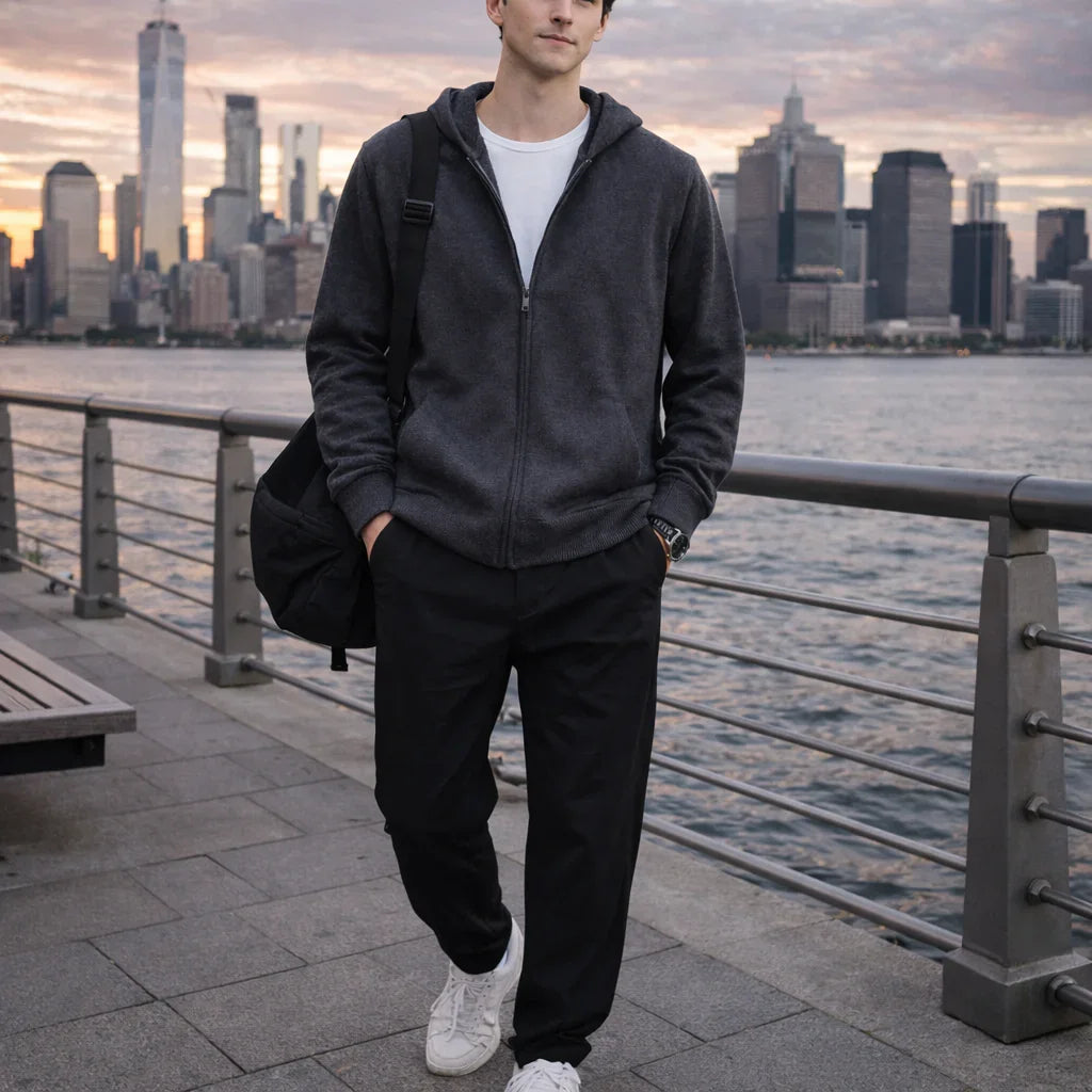Man in casual Rovelli hoodie with backpack by waterfront, city skyline at sunset