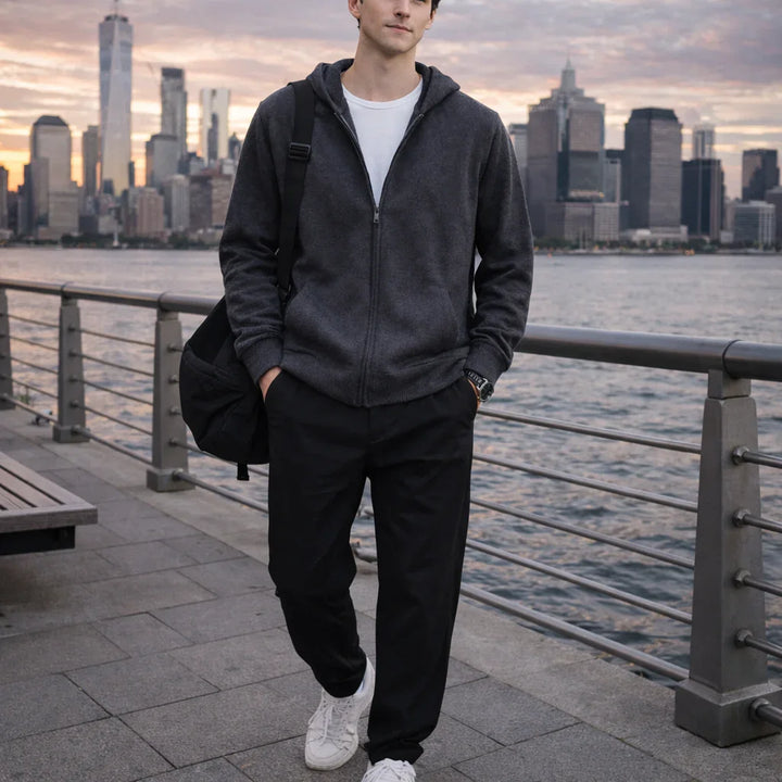 Man in casual Rovelli hoodie with backpack by waterfront, city skyline at sunset