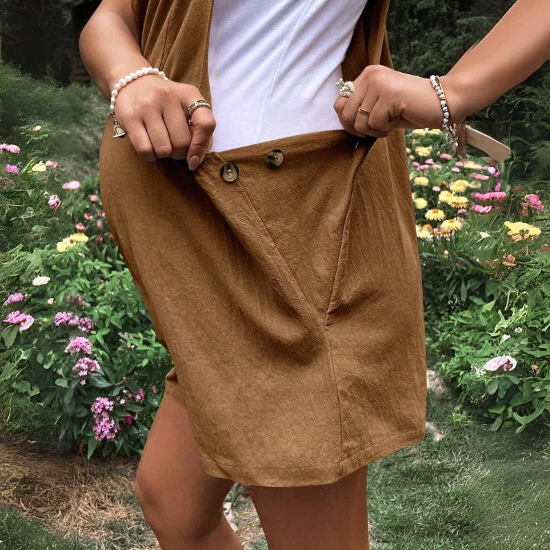 A person stands in a garden wearing comfortable maternity clothes—a brown skirt and white top—holding out the skirt with both hands to show its button details. Colorful flowers and greenery are in the background.