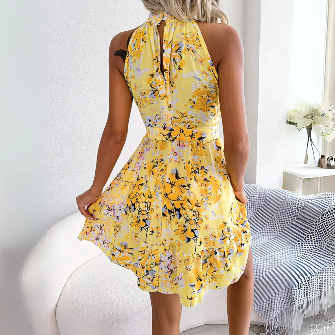 A woman in a sleeveless, yellow floral dress—perfect for a summer wardrobe—stands indoors, holding out the skirt. She faces away from the camera. The room features a white couch, checkered blanket, and a table with white flowers.