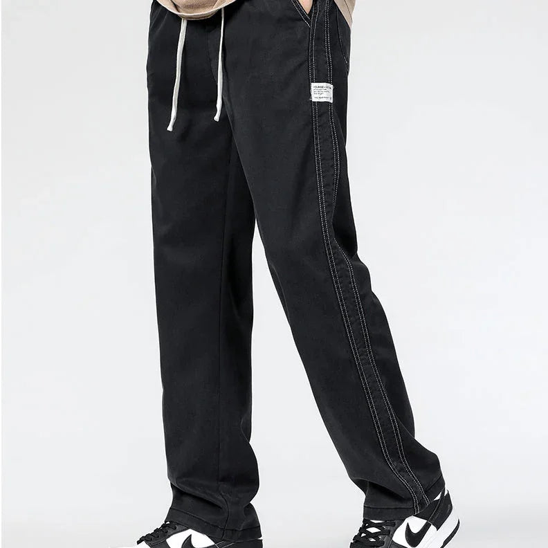 Person wearing summer loose men jeans in black with white contrast stitching and drawstrings, paired with black and white Nike sneakers, standing against a plain light background.