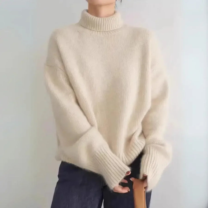 A person wearing a loose, cream-colored cashmere turtleneck sweater with ribbed cuffs and dark pants, holding a brown strap, stands against a plain light background.